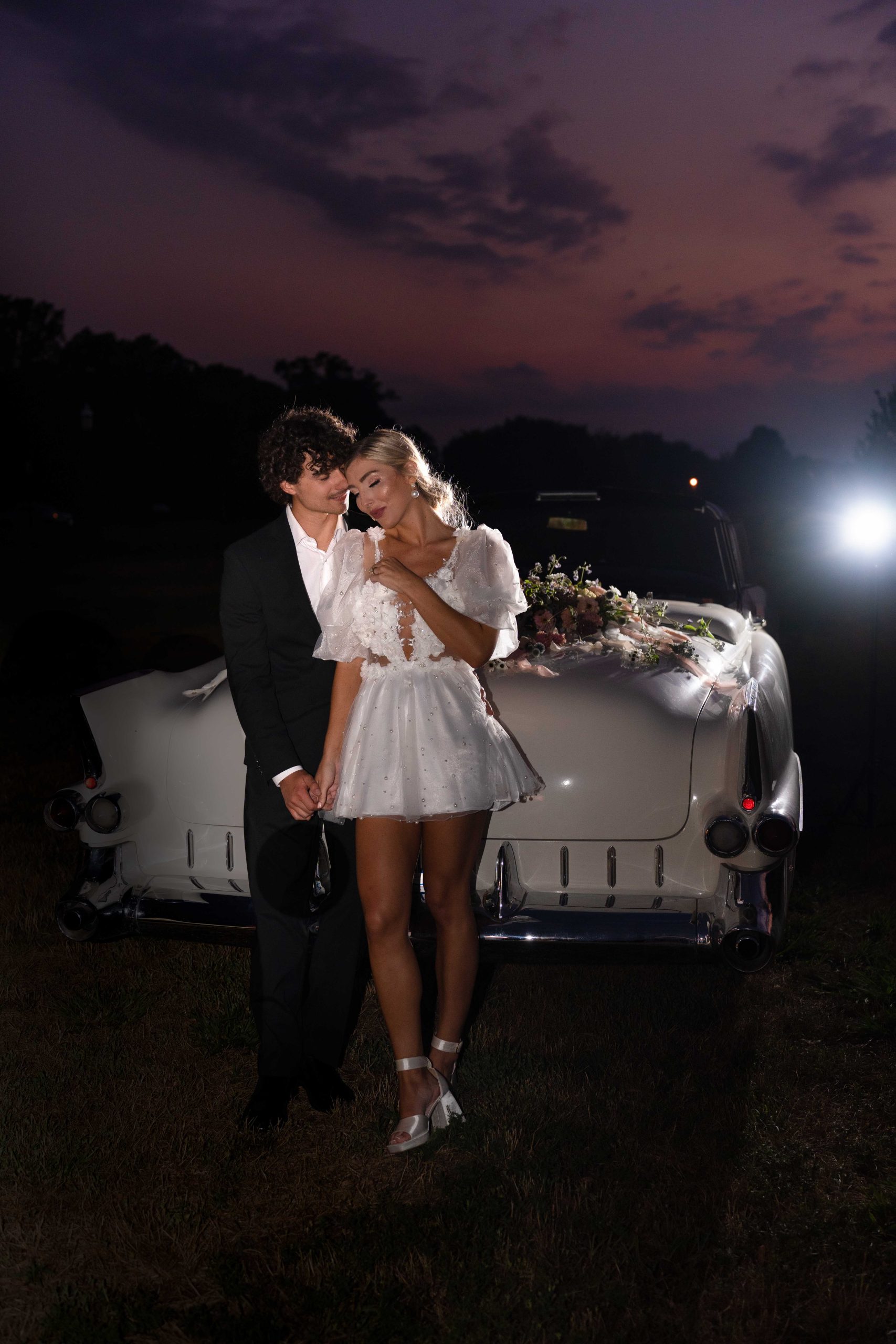 wedding couple at night leaning on a class car with sunset in the background